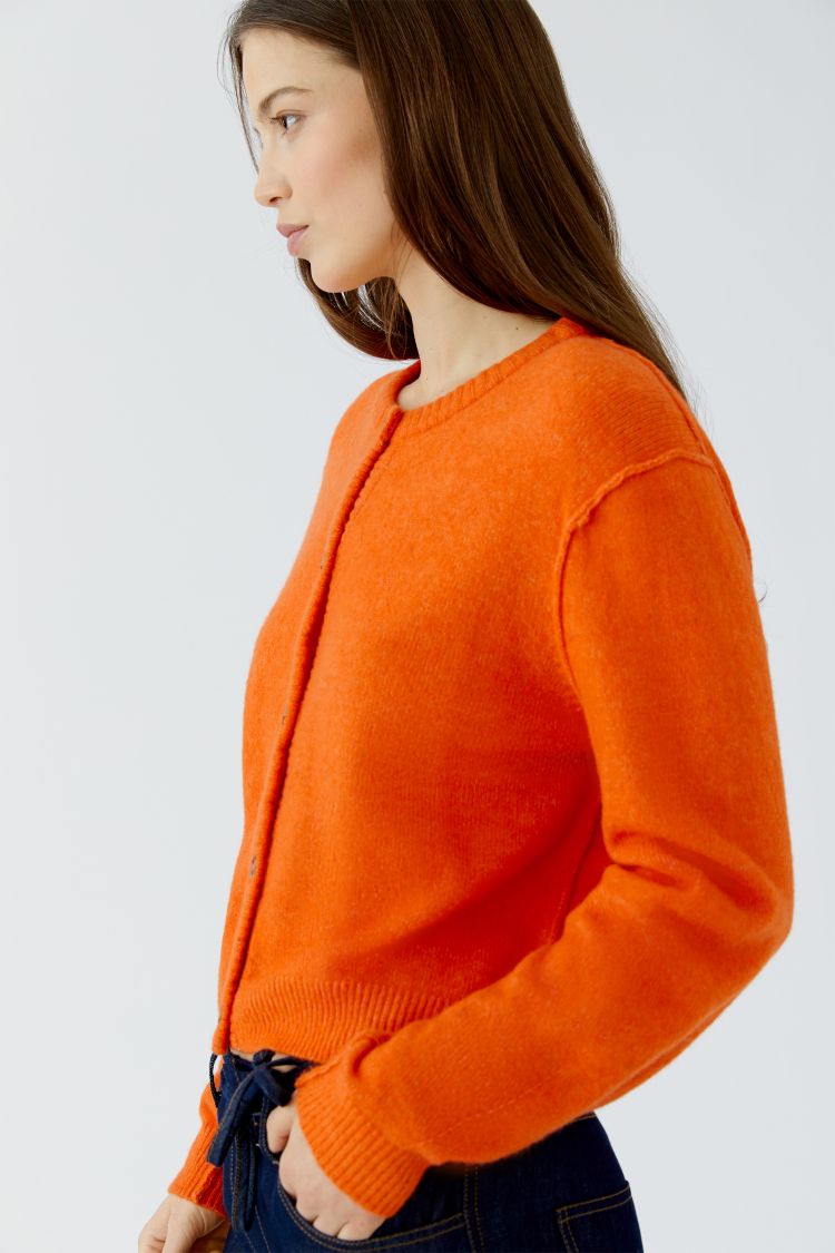 A woman wearing a bright orange knit cardigan sweater, shown in profile against a plain light gray studio background.