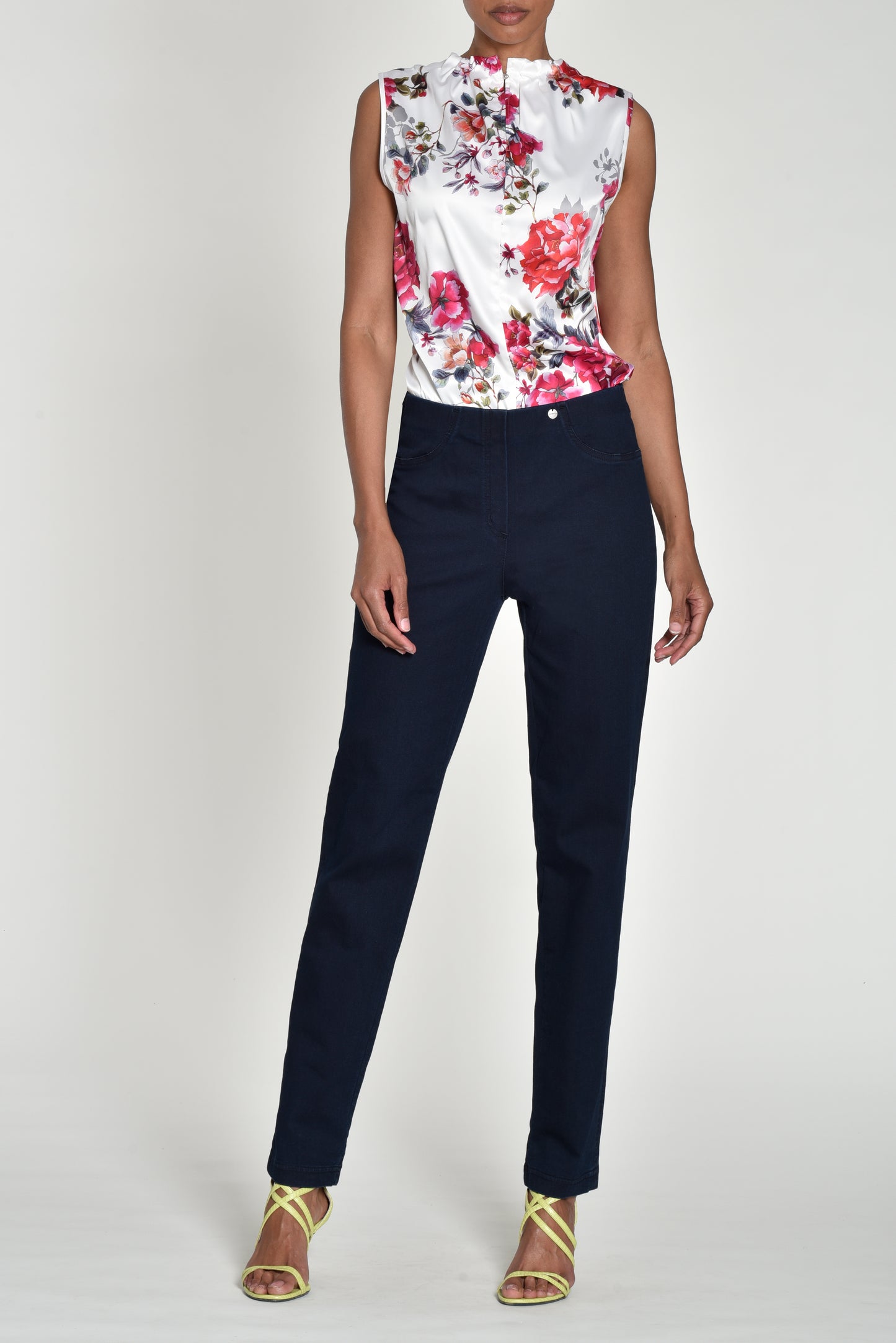 A model wearing a sleeveless white satin blouse with a vibrant red, pink, and green floral print on the front, paired with dark navy blue trousers and gold strappy high-heeled sandals, set against a plain light gray studio background.