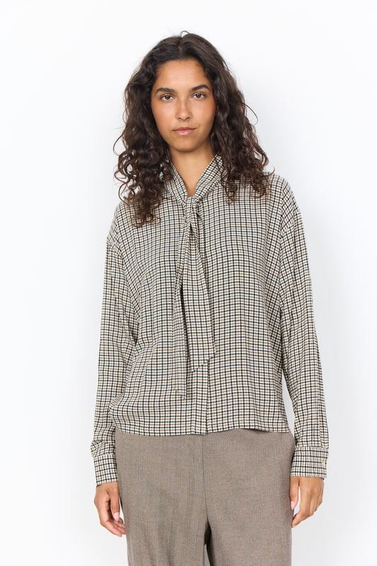 A woman with curly dark hair wearing a beige and black checkered long-sleeve blouse with a tie-neck detail, paired with gray trousers, standing against a plain white background.
