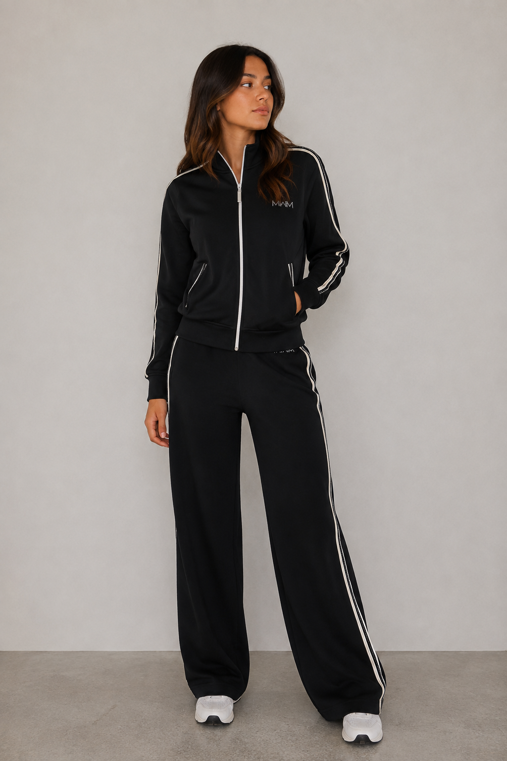 A woman modeling a black tracksuit with white stripes and a white zipper, standing against a neutral gray studio background.