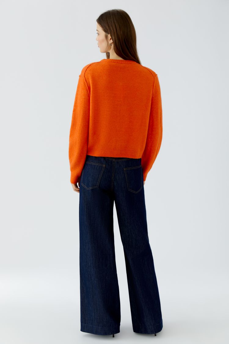 A woman shown from the back wearing a bright orange knit sweater and dark blue wide-leg jeans, photographed against a clean light gray studio background.