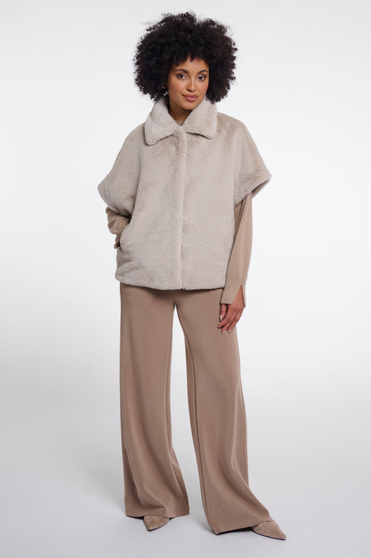 A model with an afro hairstyle wearing a light beige faux-fur cape jacket with a wide collar, paired with wide-leg taupe trousers, standing against a plain white studio background.