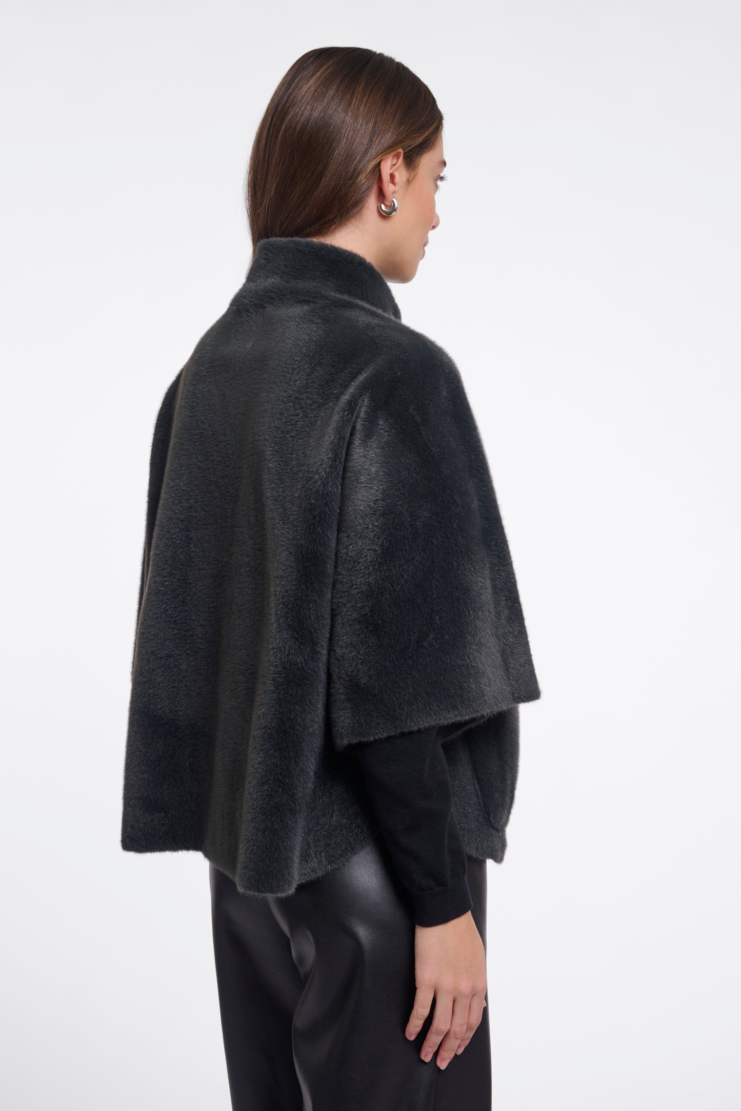 A model shown from the back wearing a dark charcoal gray faux fur cape jacket over a black long-sleeve top and black pants, against a plain white studio background.