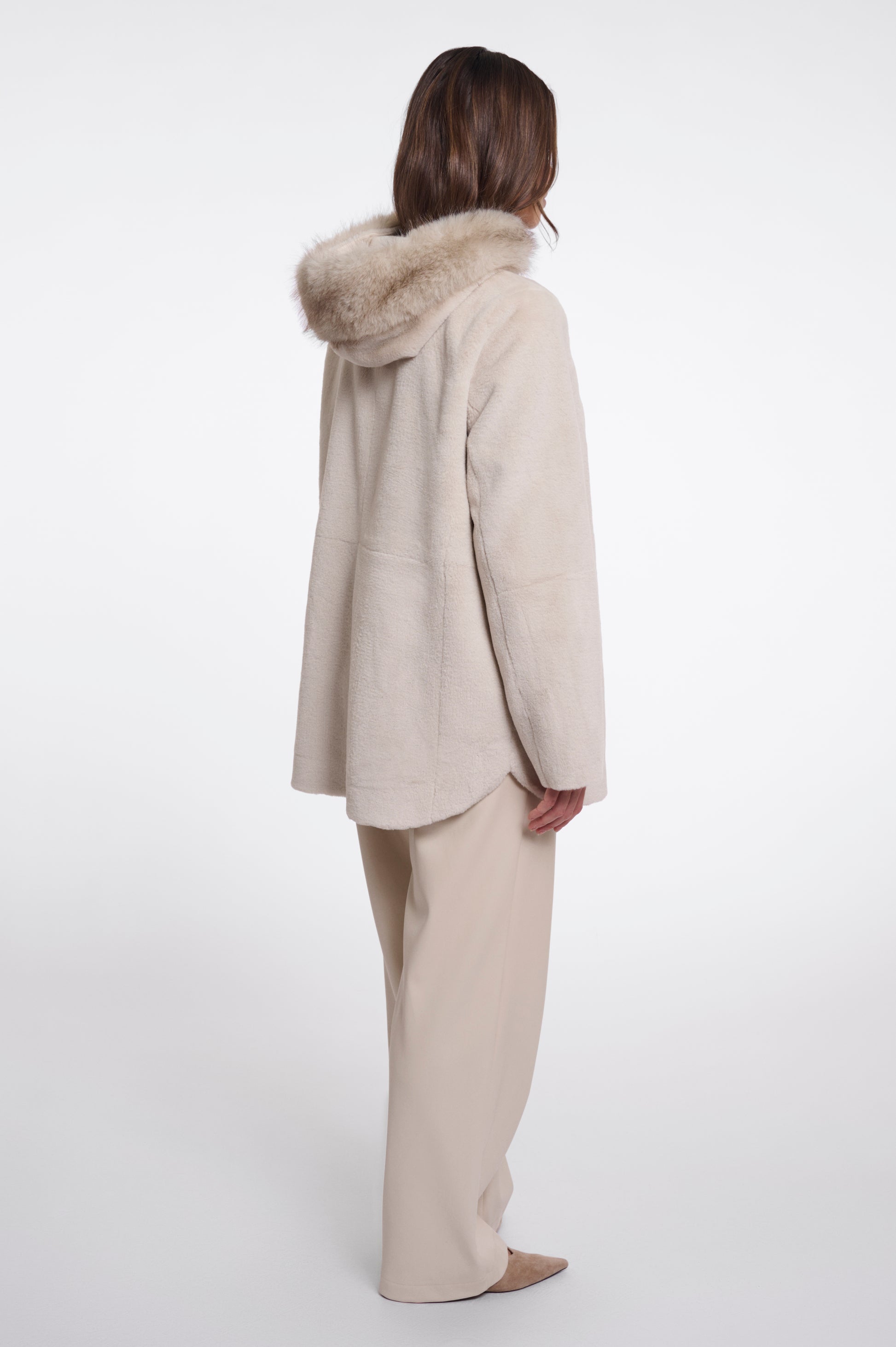 A model shown from the back wearing a light beige coat with a fluffy beige fur-trimmed hood and matching beige trousers, set against a plain white studio background.