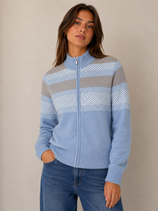A woman wearing a light blue zip-up knit sweater with beige and white patterned horizontal stripes, paired with blue jeans, standing against a neutral beige background.